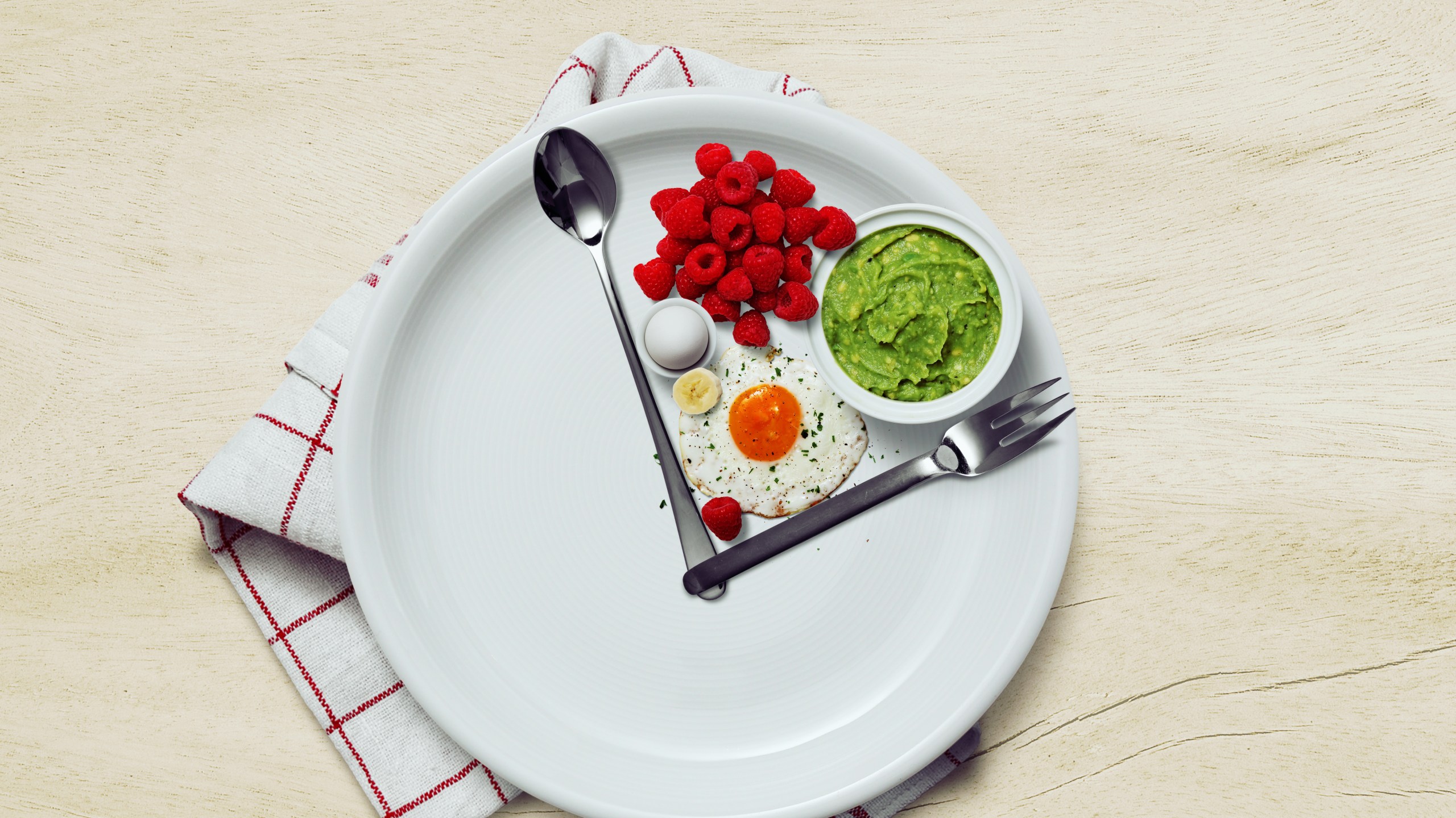 A white plate with raspberries, an egg, a small bowl of avocado and a spoon and knife.