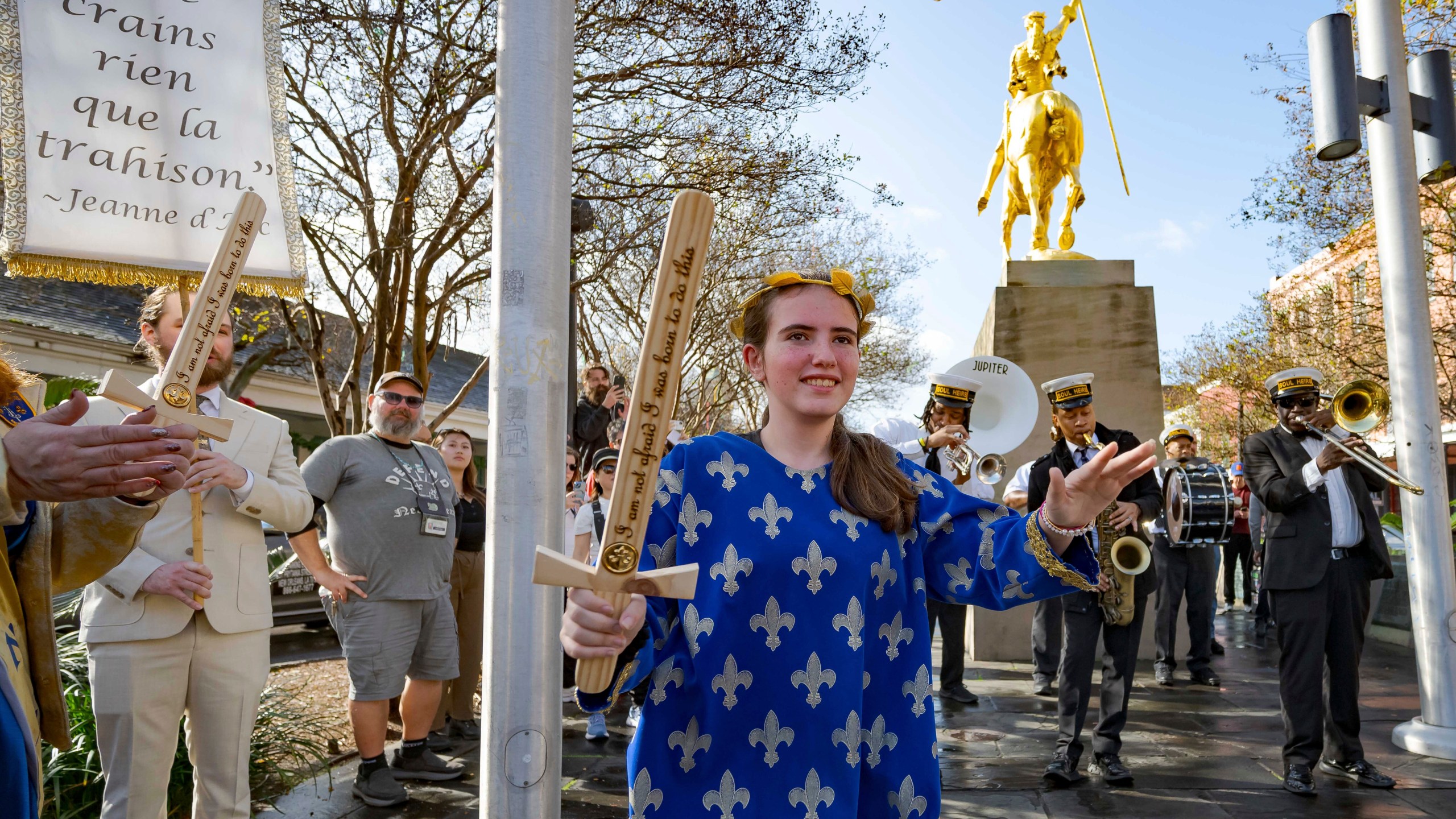 A procession of Krewe de Jeanne d'Arc represented by 2025 Maid of Honor Life Sacco, center, and the Soul Heirs Brass Band dances by a golden statue of St. Joan of Arc in the French Quarter after leaving St. Louis Cathedral in New Orleans, Sunday, Jan. 5, 2025, after a mass honoring victims of the New Year's Day deadly truck attack and shooting not far from the cathedral in the French Quarter. (AP Photo/Matthew Hinton)