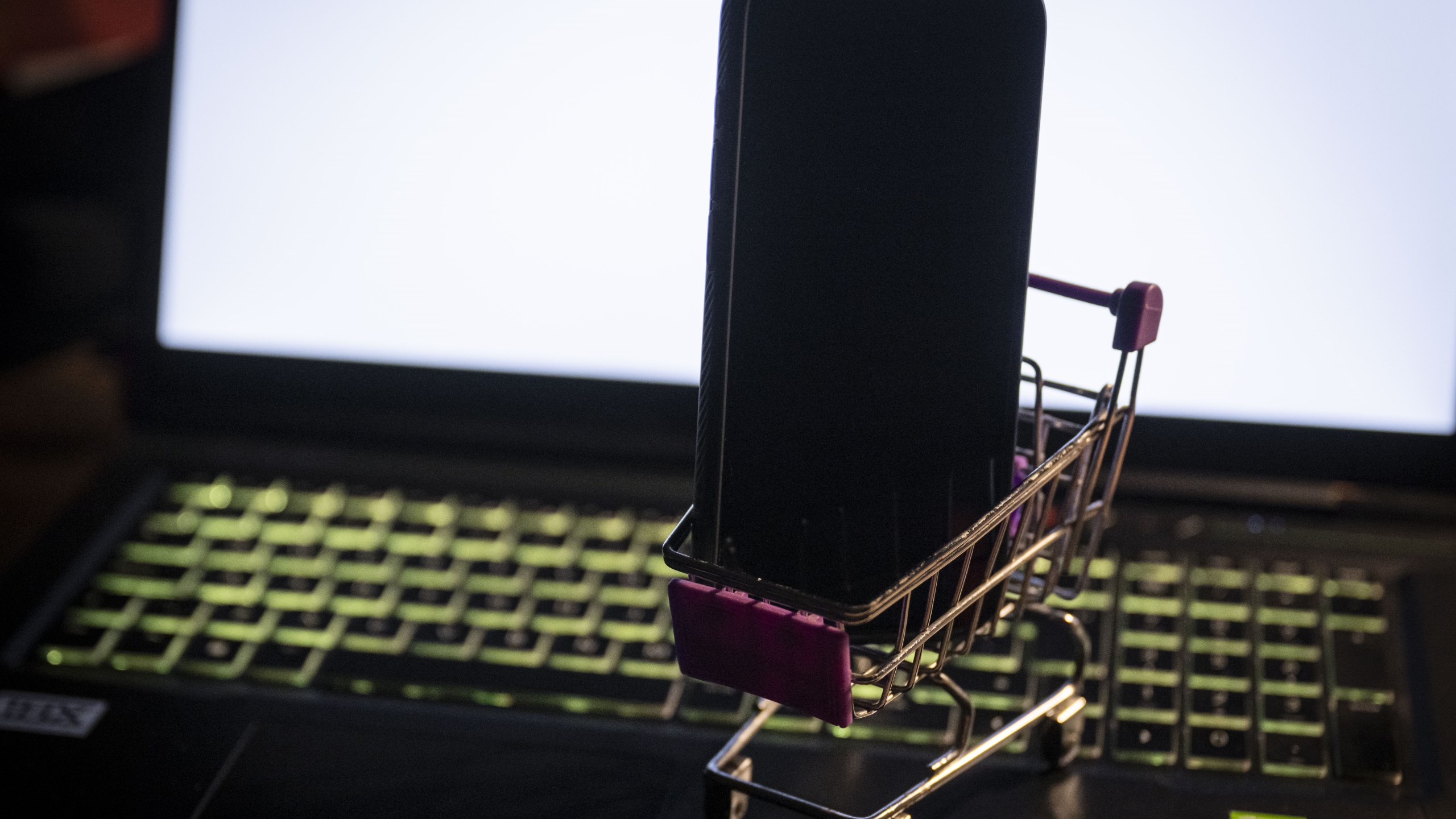 A miniature shopping cart with a phone is seen on a laptop with its screen displaying an online shopping website.