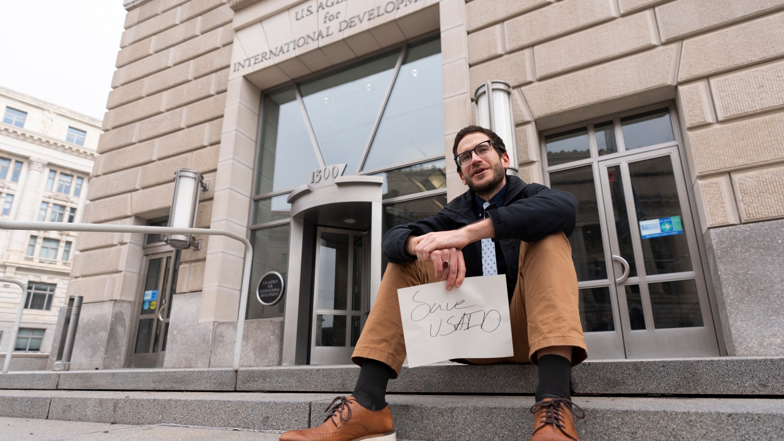 A United States Agency for International Development, or USAID, contract worker sits in front of the USAID office with a message written on a piece of paper, Monday, Feb. 3, 2025, in Washington (AP Photo/Manuel Balce Ceneta)