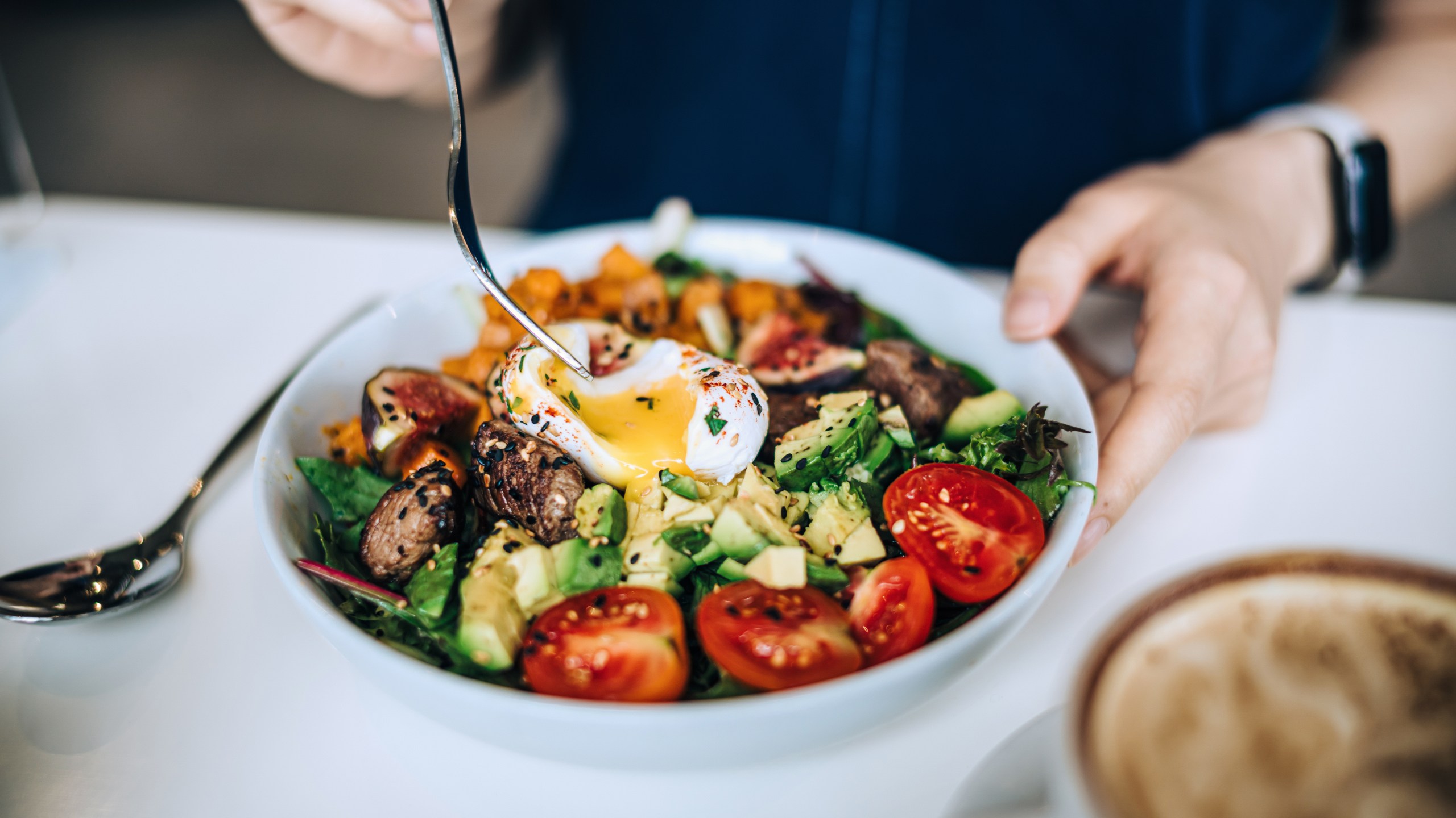 Close up shot of a woman eating a dish of fresh beef cobb salad with a soft boiled egg and coffee at a cafe table.
