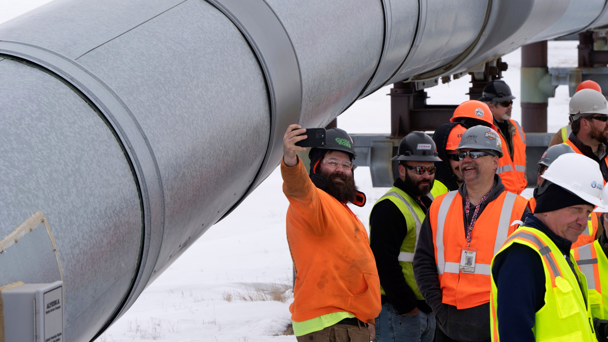 A worker poses for a photo with start of the Trans-Alaska Pipeline in the background after a news conference at the Pump Station 1 on Monday, June 2, 2025, located near Deadhorse, Alaska, on the state's prodigious North Slope. (AP Photo/Jenny Kane)