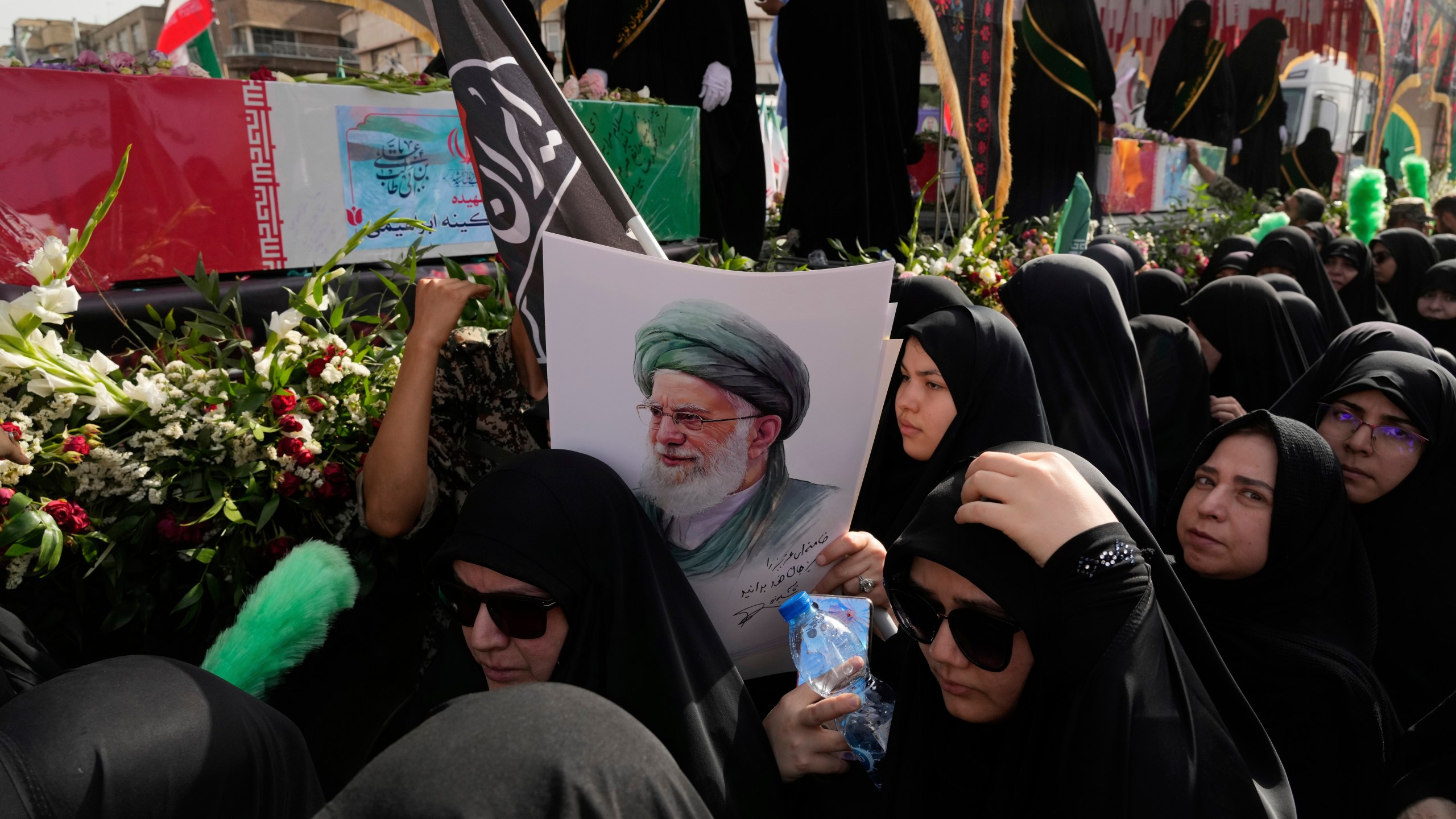 Mourners gather around the flag-draped coffins of the Iranian armed forces generals, nuclear scientists and their family members who were killed in Israeli strikes, during their funeral ceremony as one of them holds a poster of the Supreme Leader Ayatollah Ali Khamenei, in Tehran, Iran, Saturday, June 28, 2025. (AP Photo/Vahid Salemi)