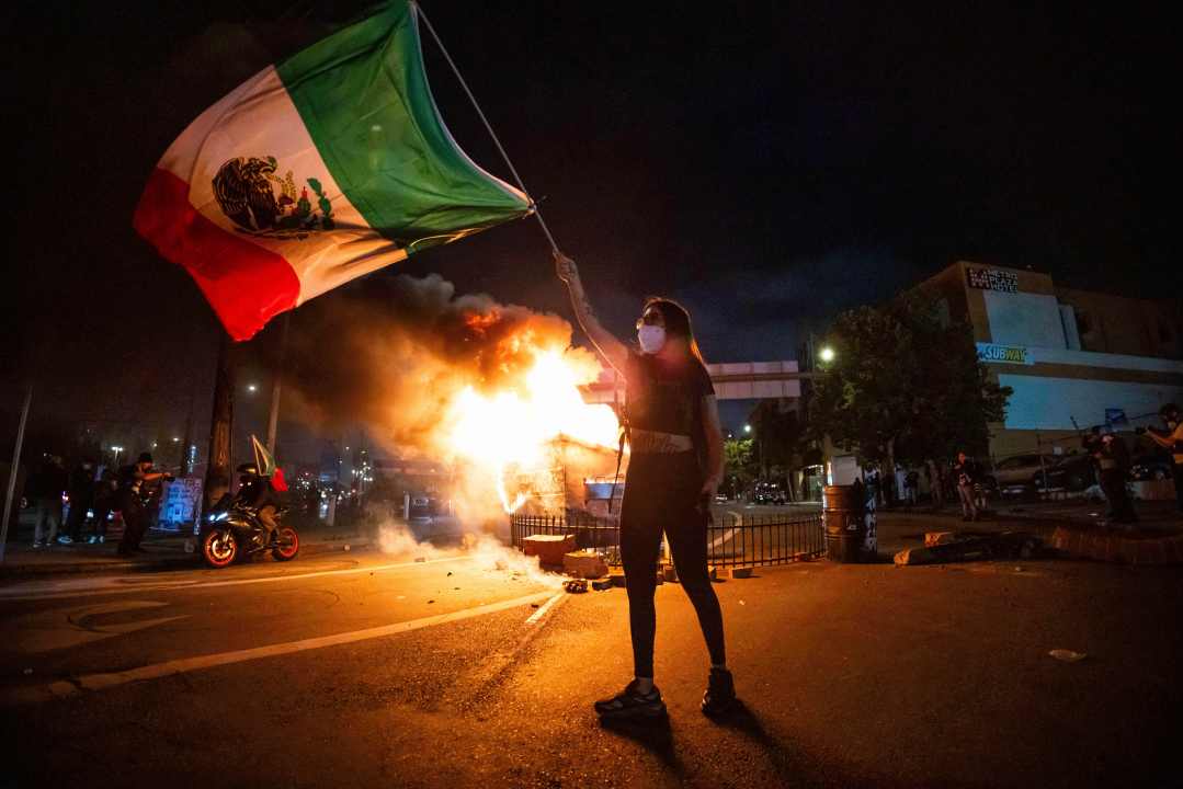A woman waves the Mexican flag as flames erupt from a burning dumpster