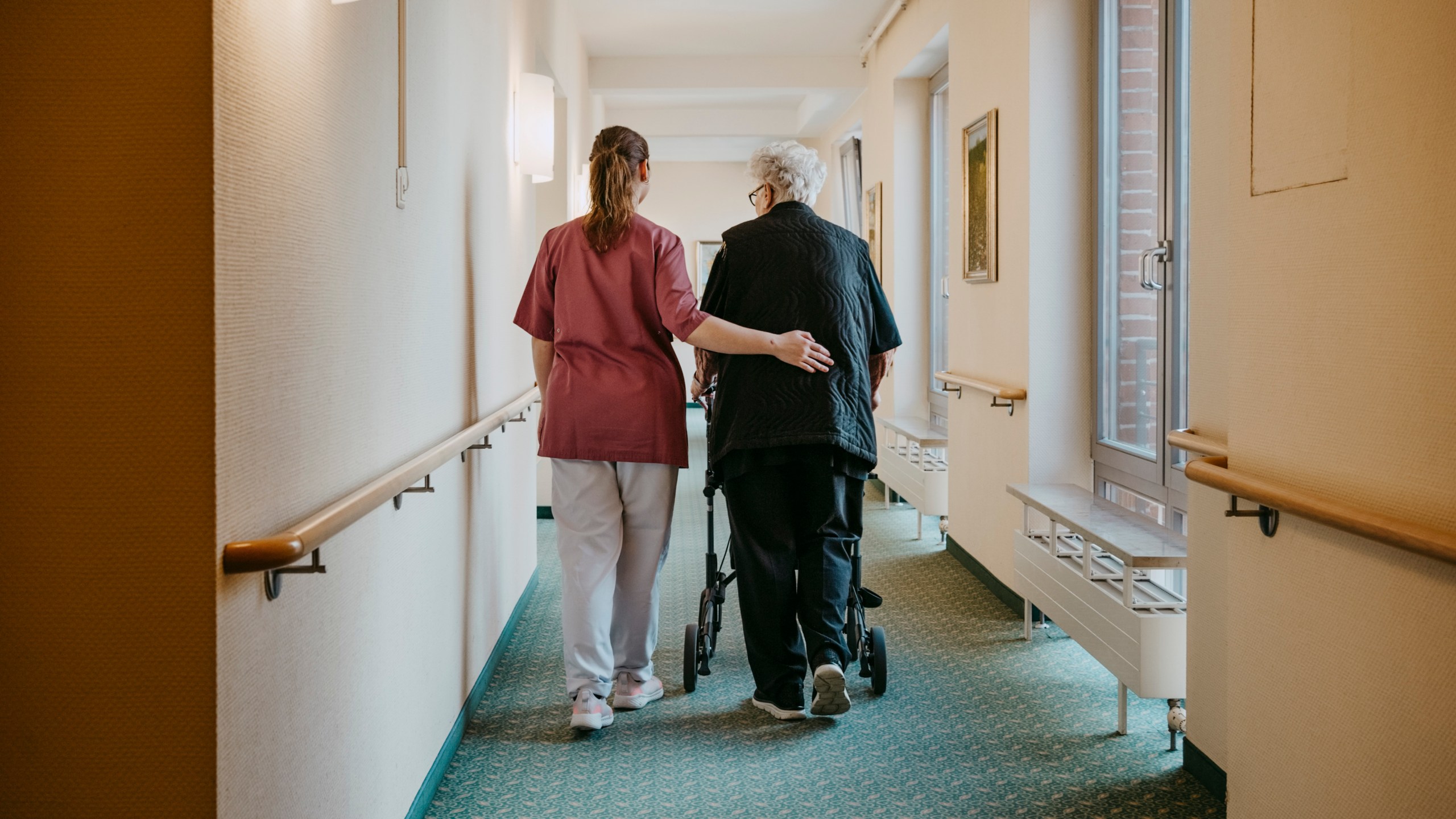 A caregiver assisting a senior woman walking with mobility walker in a nursing home corridor.