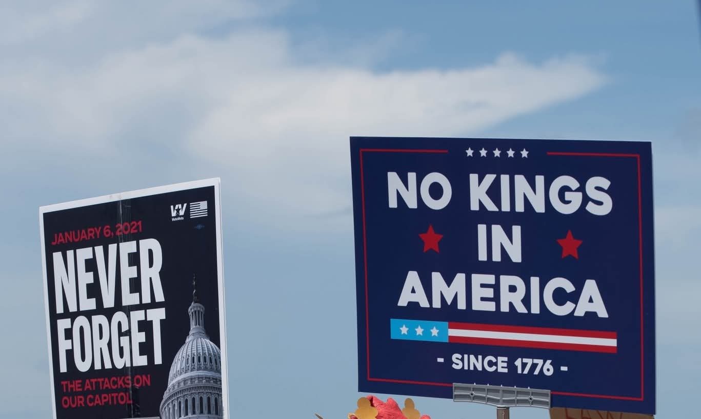 protest signs reading NEVER FORGET and NO KINGS IN AMERICA