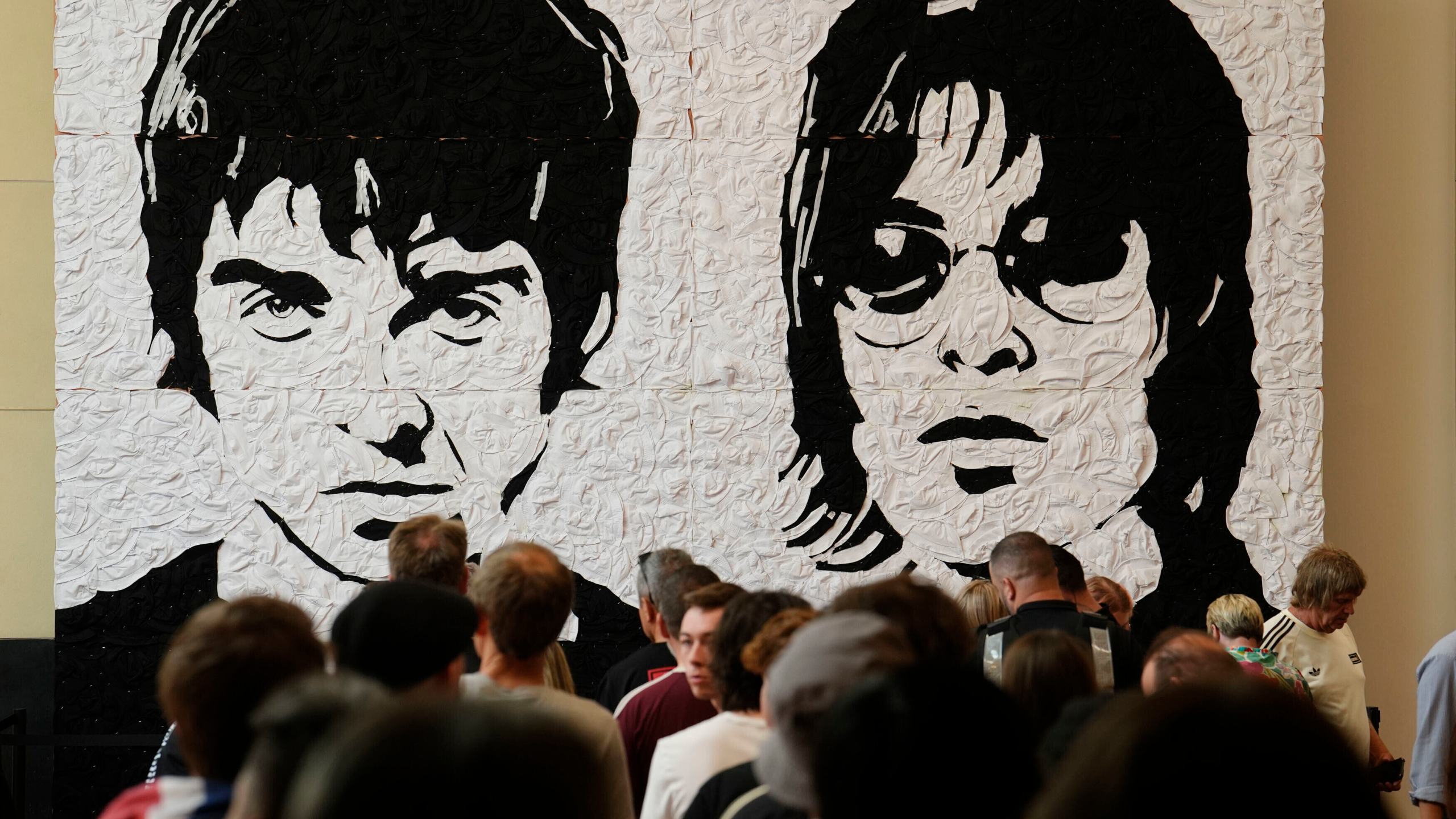 Fans de Oasis se toman fotos frente al Wonder Wall, un mural del artista galés Nathan Wyburn hecho con 3.000 sombreros de pescador blancos y negros que representan a Liam y Noel Gallagher, en el centro comercial St David's en Cardiff, antes del primer espectáculo de su tan esperada gira de reunión que comienza en Gales, el viernes 4 de julio de 2025. (Jordan Pettitt/PA via AP)