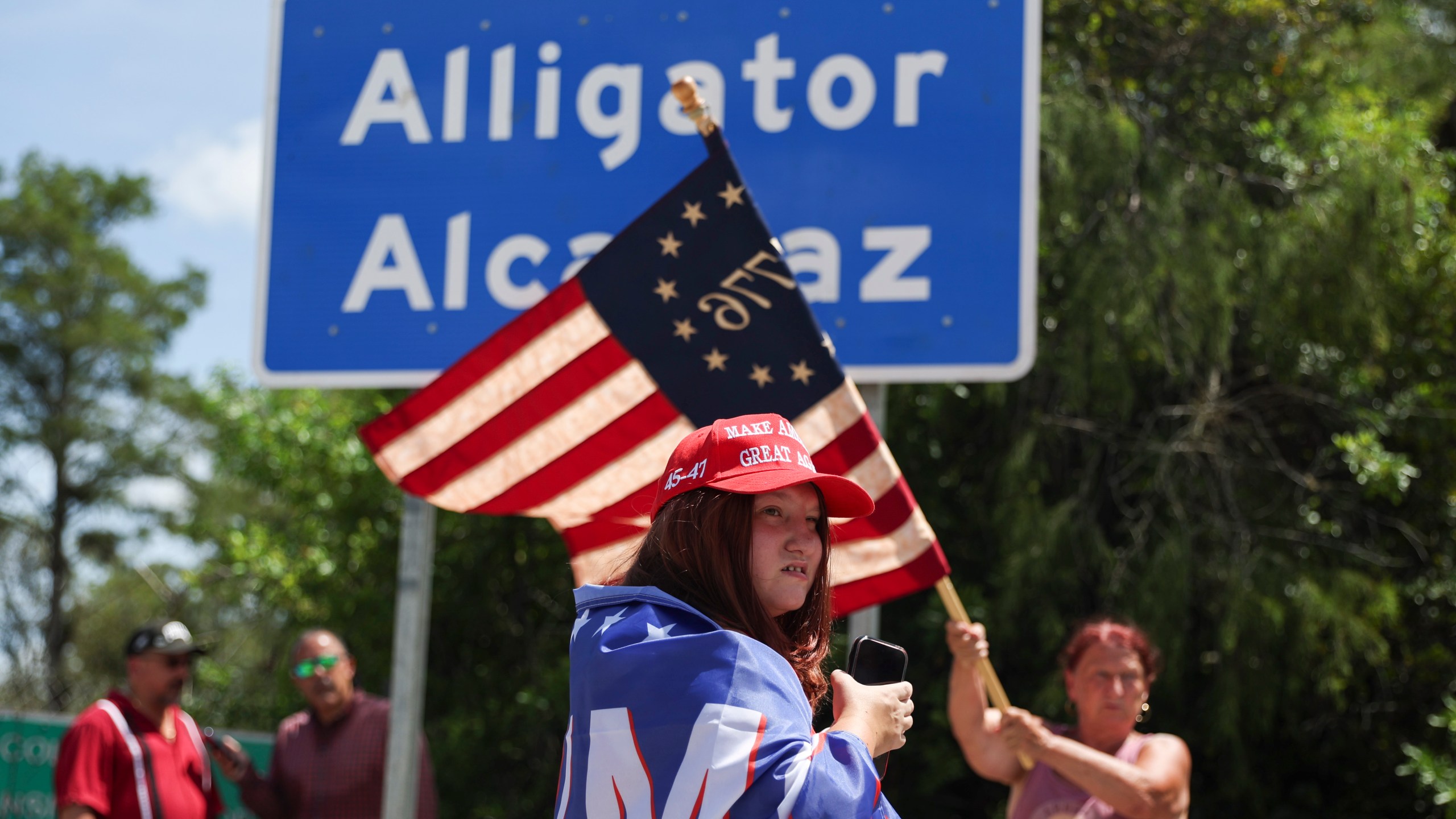Dianne Mourer waves an American flag as Rana Mourer stands in front of a sign reading "Alligator Alcatraz" outside the Dade-Collier Training and Transition Facility, Saturday, July 12, 2025, in Ochopee, Fla. (AP Photo/Alexandra Rodriguez)