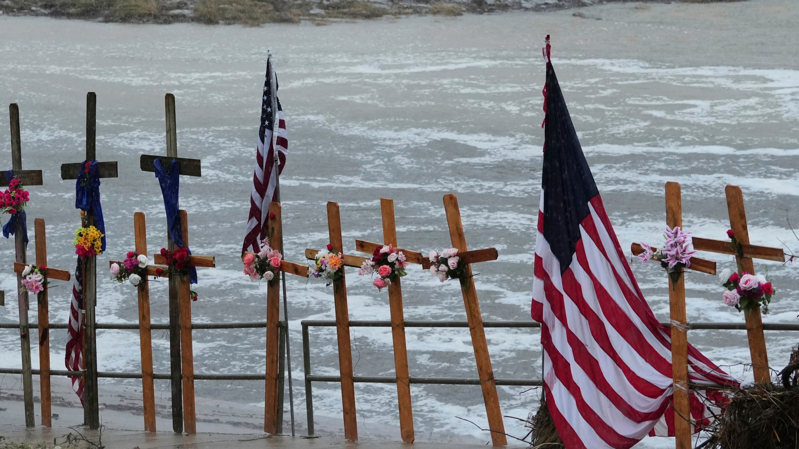 rain falls over a makeshift memorial, crosses and flags next to a river