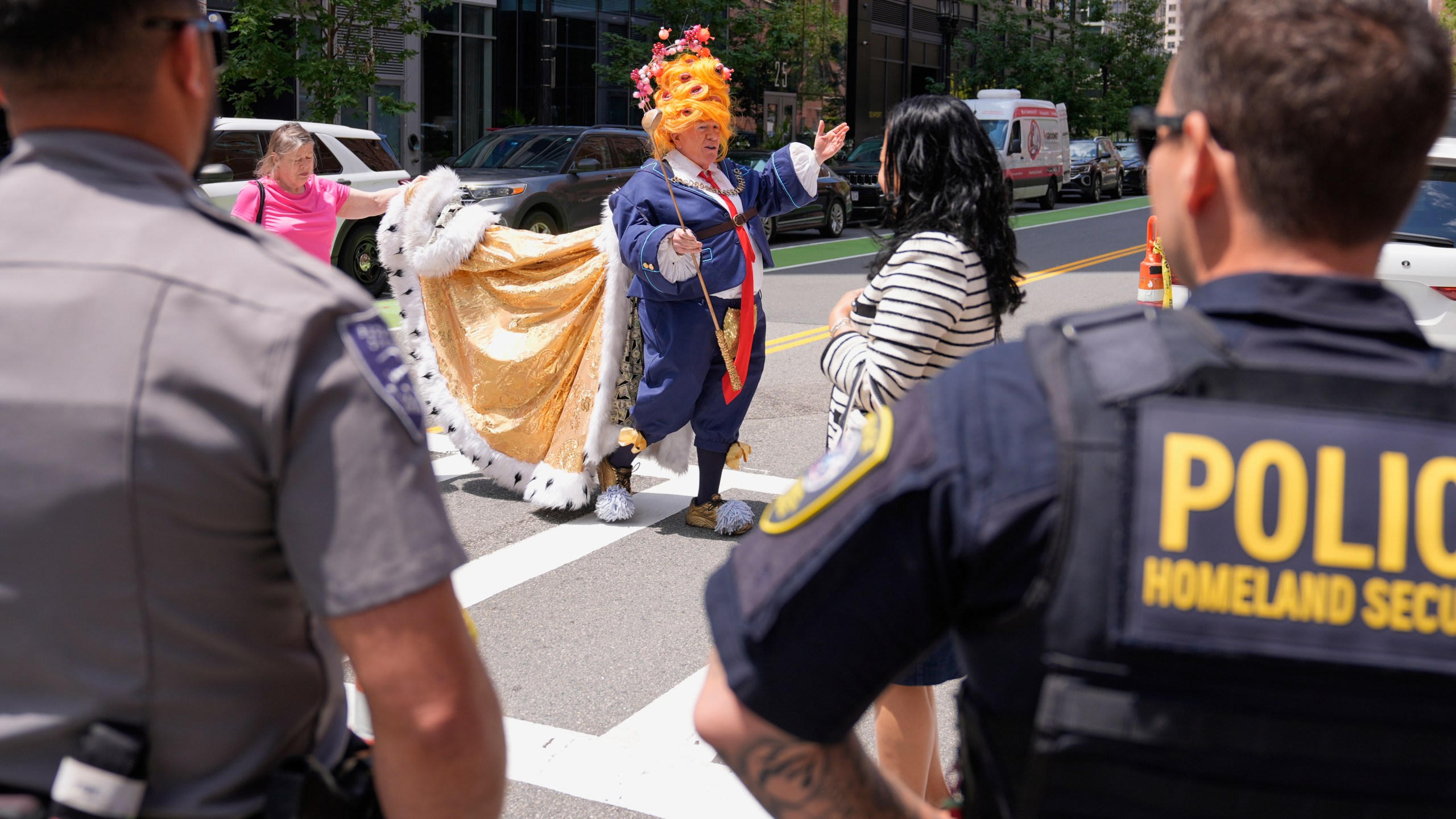 Law enforcement watch as an actor dressed in a costume, who identified himself as "the real mad king", walks toward protestors outside the Moakley Federal Courthouse, where Harvard University appeared to challenge $2.6 billion in funding cuts by the Trump administration, Monday, July 21, 2025, in Boston. (AP Photo/Charles Krupa)