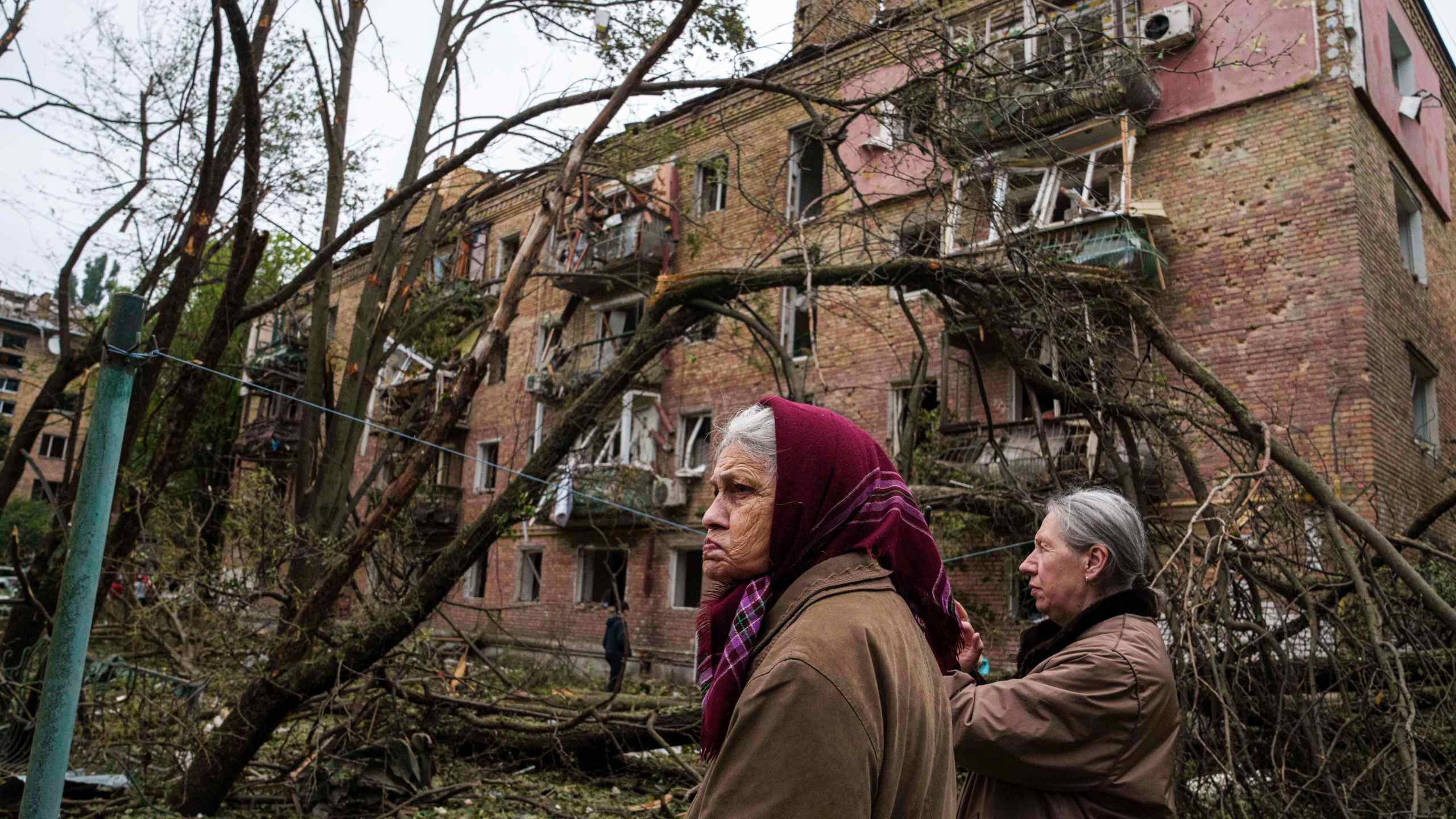 women stand in front of a residential building heavily damaged by bombs