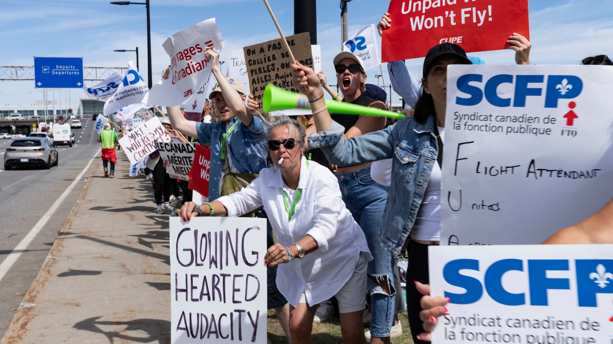 protesters gather holding signs reading GLOWING HEARTED AUDACITY and UNPAID WORK WON'T FLY
