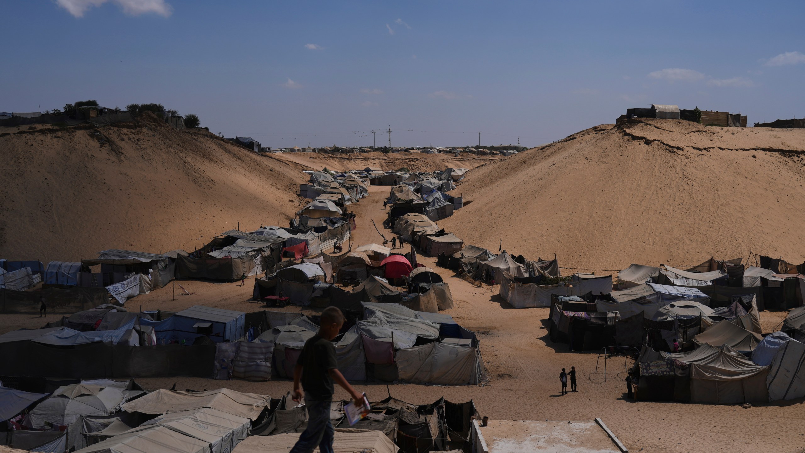 Displaced Palestinians walks through a tent camp in Muwasi, an area that Israel has designated as a safe zone, in Khan Younis southern Gaza Strip, Sunday, Sept. 21, 2025. (AP Photo/Jehad Alshrafi)