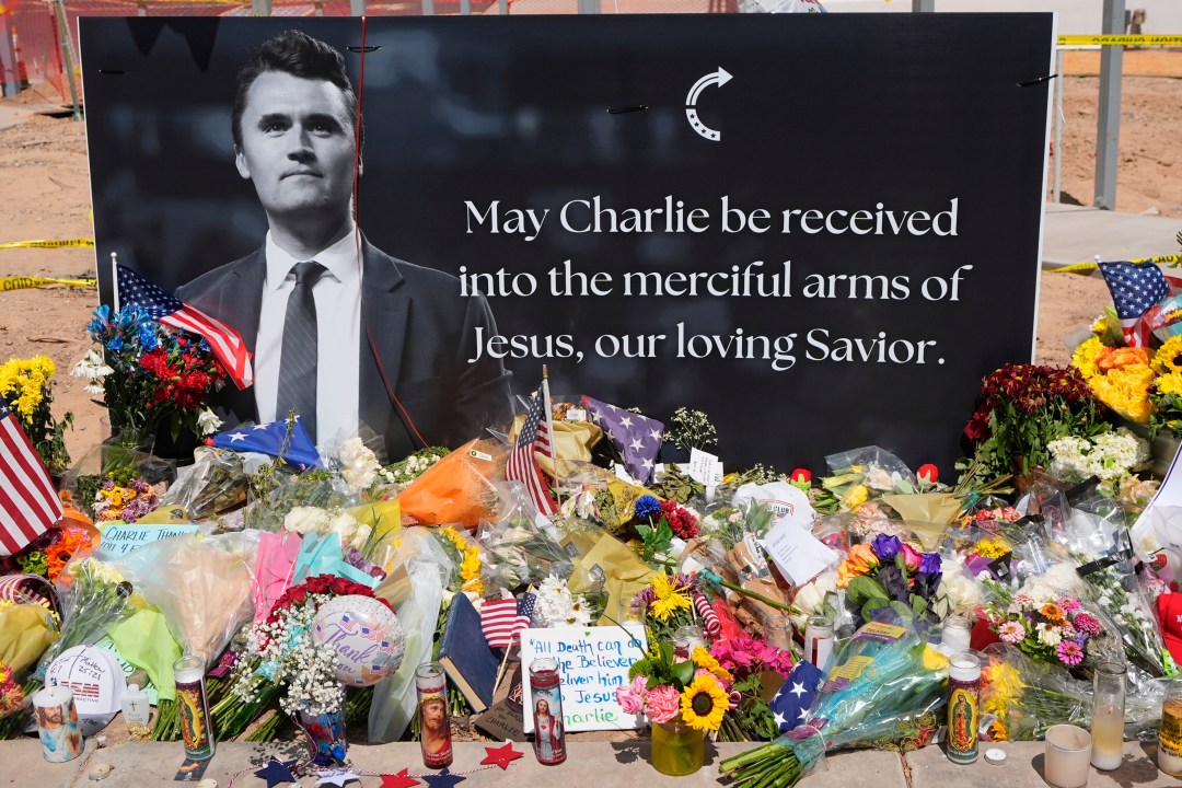 a memorial for Charlie Kirk with flowers and a sign reading MAY CHARLIE BE RECEIVED INTO THE MERCIFUL ARMS OF JESUS, OUR LOVING SAVIOR