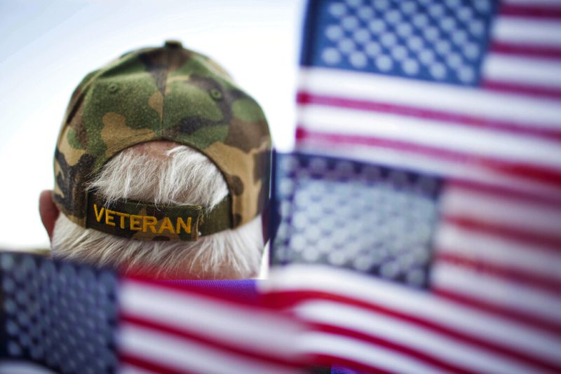 Photo of the back of a man's head with a hat that says "veterans" surrounded by American flags