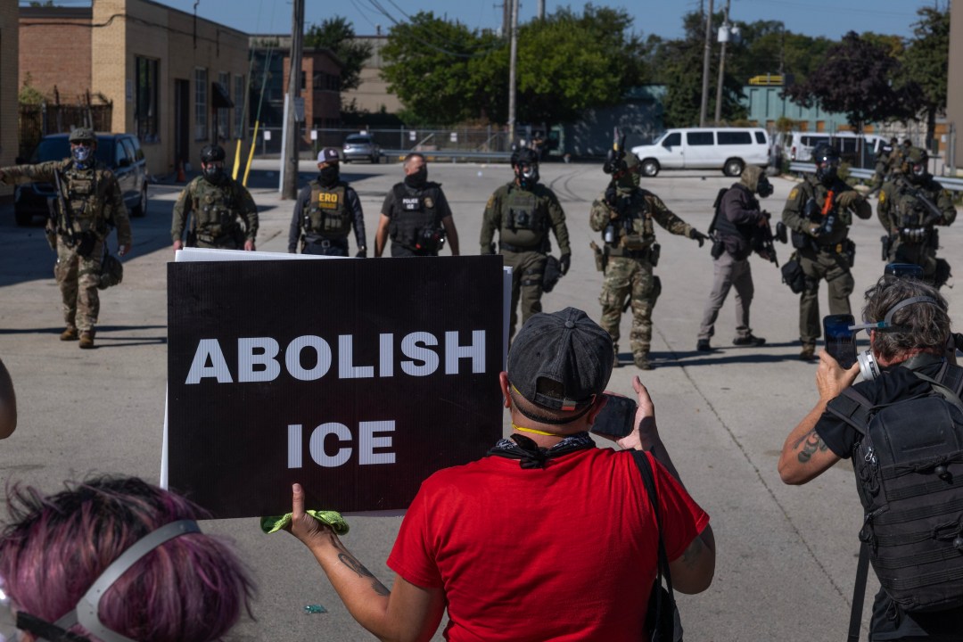 Protesters gather in front of ICE officers. One holds a sign reading ABOLISH ICE