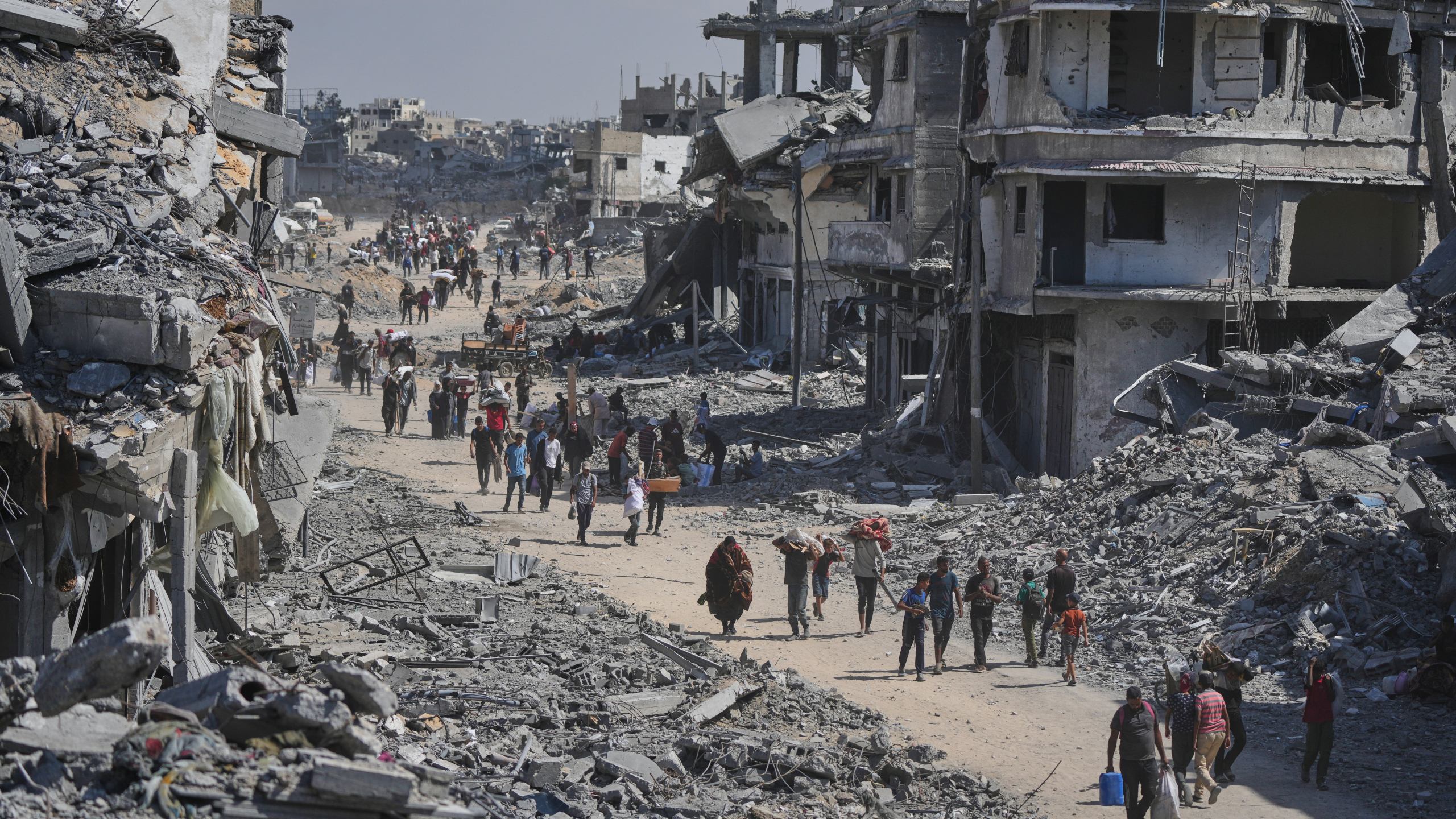 Displaced Palestinians walk with their belongings past destroyed buildings as they return to their homes in Khan Younis, southern Gaza Strip, Friday, Oct. 10, 2025, after Israel and Hamas agreed to a pause in their war and the release of the remaining hostages. (AP Photo/Jehad Alshrafi)
