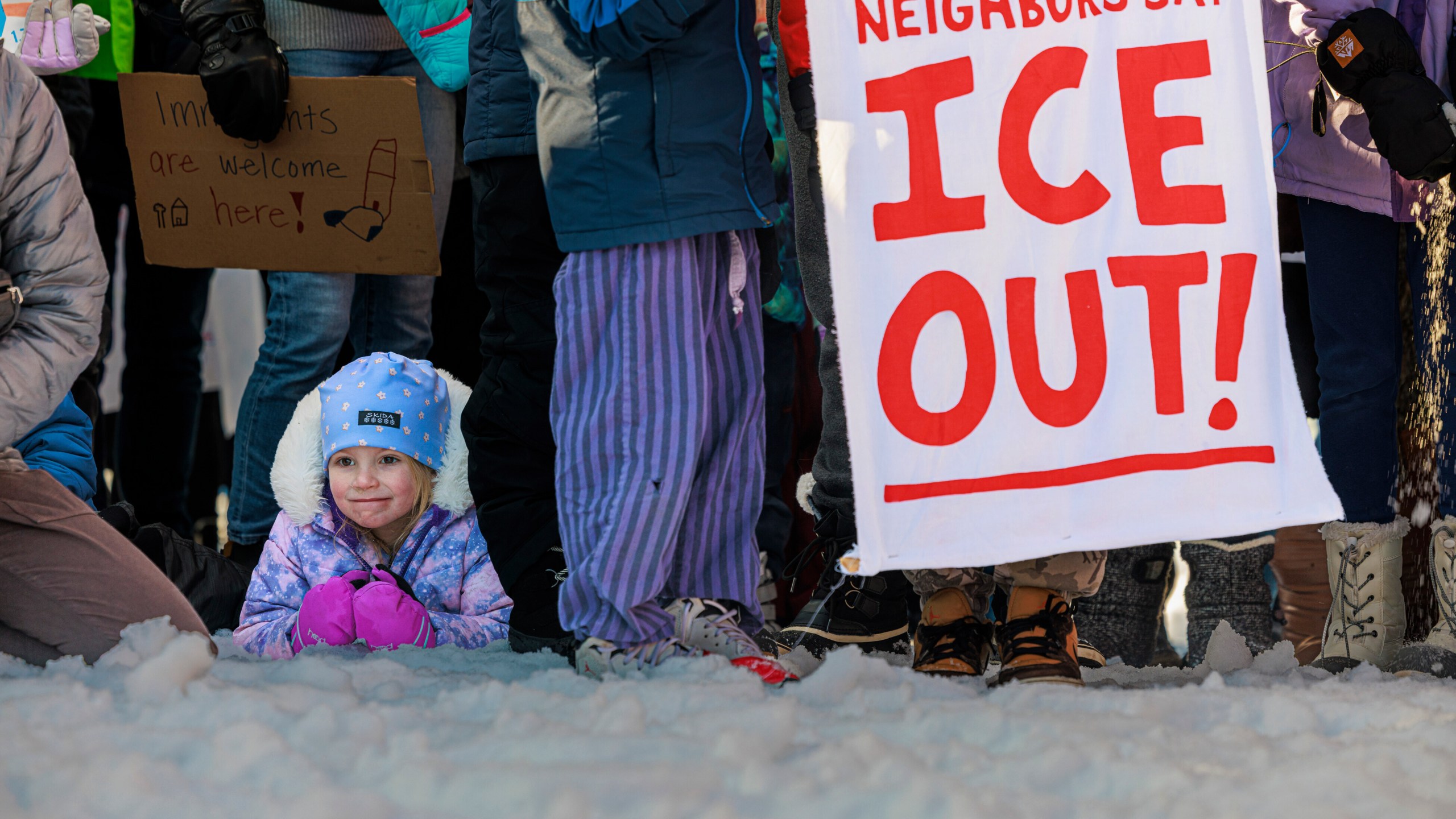 A child sits in the snow as demonstrators hold signs during a news conference at Lake Hiawatha Park in Minneapolis, on Friday, Jan. 9, 2026, demanding Immigration and Customs Enforcement be kept out of schools and Minnesota following the killing of 37-year-old mother Renee Good by federal agents earlier on Wednesday. (Kerem Yücel/Minnesota Public Radio via AP)
