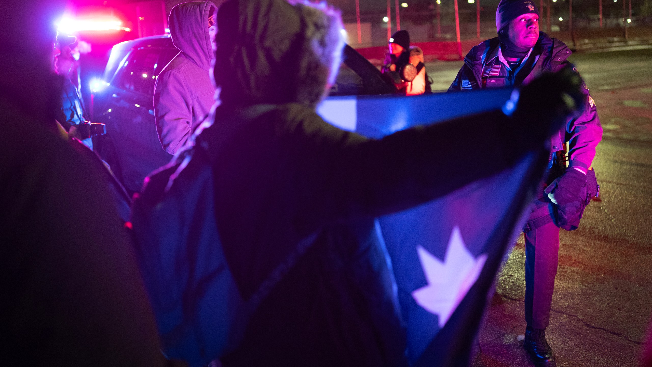 deputy stands near protester with flag, emergency lights flashing